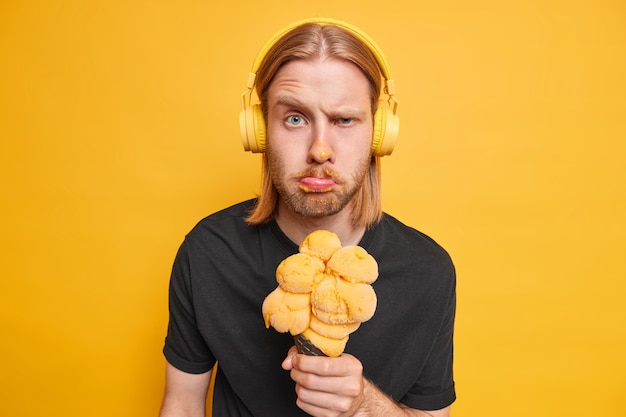 Free photo displeased redhead man frowns face makes unhappy grimace raises eyebrows has dirty face while eating ice cream wears wireless stereo headphones for listening music isolated over yellow  wall