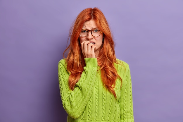 Displeased woman bites finger nails frowns face looks unhappily feels nervous because of something.