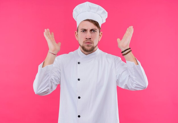 A displeased young bearded chef man in white uniform raising both hands up on a pink wall