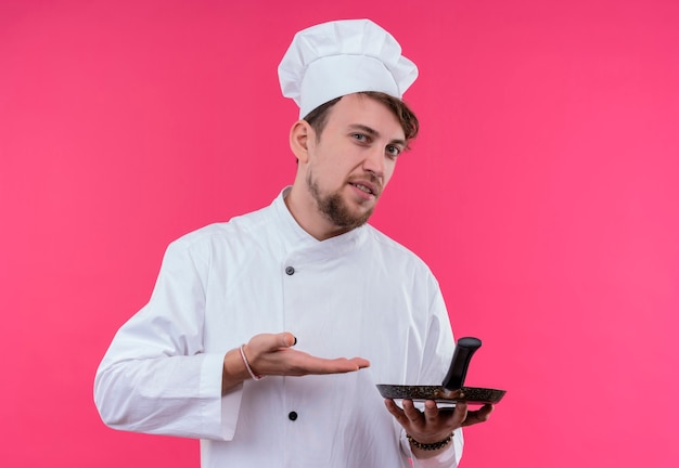 A displeased young bearded chef man in white uniform showing frying pan while looking on a pink wall