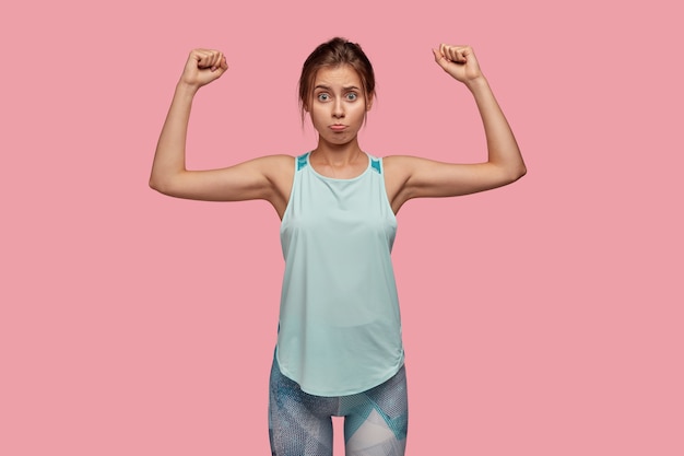 Free Photo displeased young caucasian blue eyed woman with unhappy sullen expression, has nice perfect figure raises hands and shows muscles, wears activewear, isolated over pink wall, feels weak