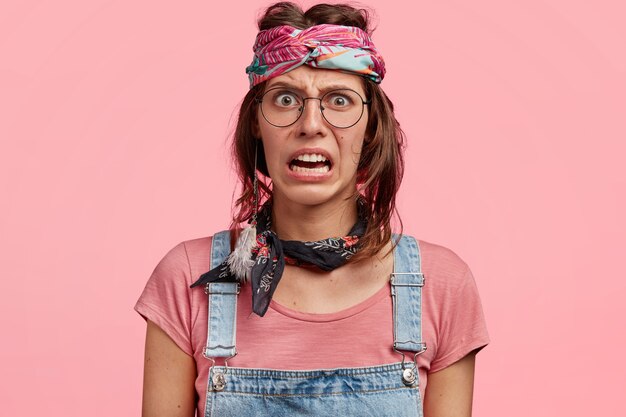 Displeased young female hippie with discontent expression, curves lips, sees something negative, dressed in casual t-shirt, overalls and headband, isolated over pink wall. Negative emotions