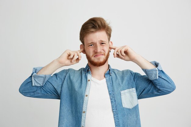 Displeased young handsome man with beard frowning closing ears.