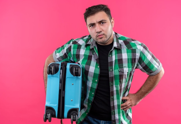 Free Photo displeased young traveler man in checked shirt holding suitcase with angry face standing over pink wall