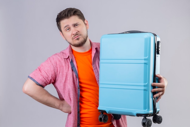 Free Photo displeased young traveler man holding blue suitcase with skeptic expression on face standing over white wall