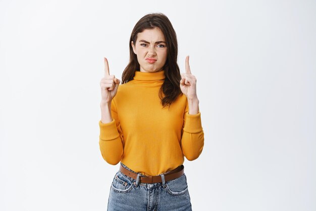 Displeased young woman express dislike pointing fingers up at something bad grimacing and squinting disappointed standing against white background