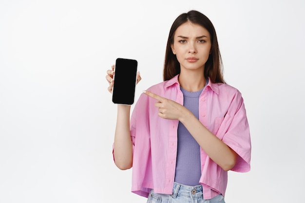 Dissapointed teenage girl pointing at smartphone screen frowning as expressing dislike displeased emotion standing over white background