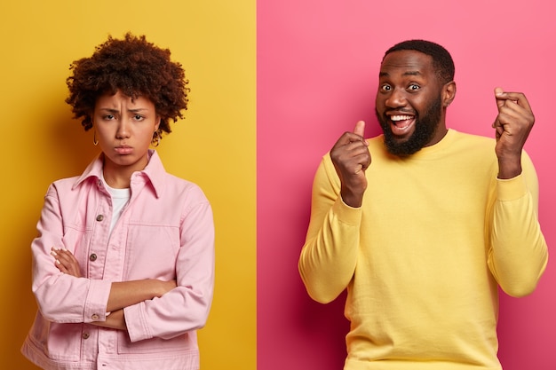 Free Photo dissatisfied african american woman stands with arms folded, displeased after quarrel with husband, triumphing dark skinned man raises both arms. ethnic couple stand over two colored wall