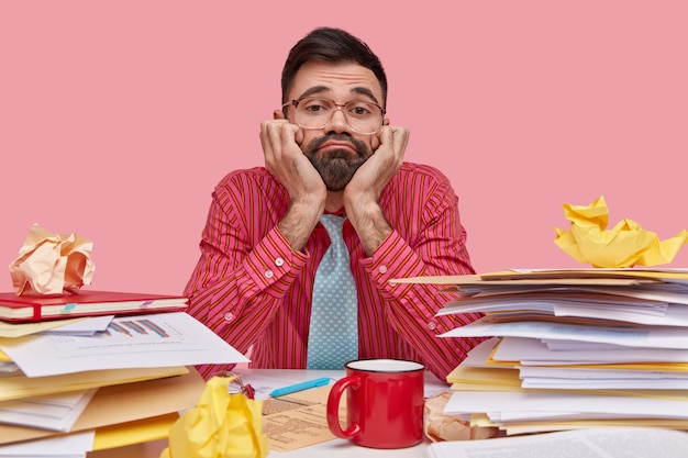 Free photo dissatisfied bearded man keeps hands under chin, looks through spectacles with apathy, wears formal clothes, surrounded with stack of papers