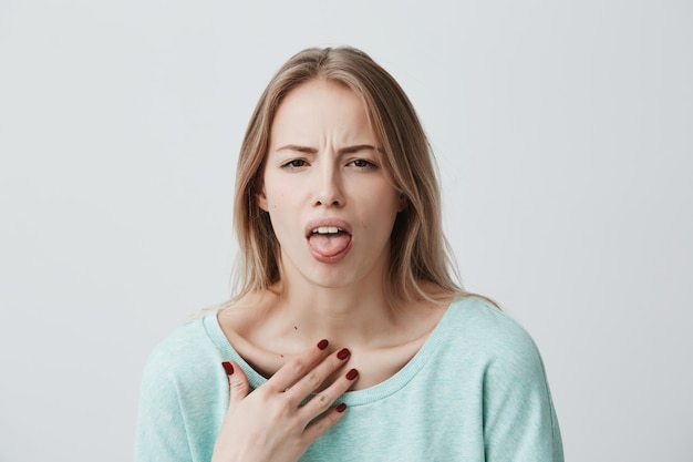 Free photo dissatisfied blonde female model frowns face, has disgusting expression, shows tongue, expresses non-compliance, irritated with somebody, rejects do something. people and negative facial expressions