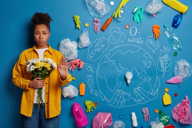 Free photo dissatisfied curly haired afro woman makes stop gesture, holds flowers in hands, asks humanity to stop and think about cleaning nature