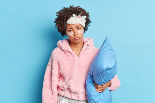 Dissatisfied curly haired young Afro American woman wants to sleep holds pillow prepares to go to bed