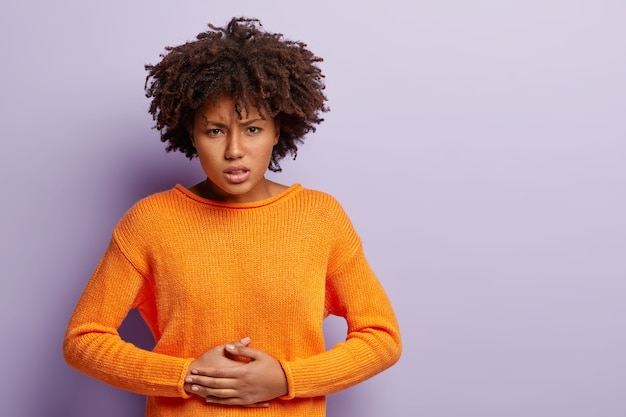 Free photo dissatisfied dark skinned female keeps hands on stomach, suffers from pain, has menstruation, wears orange jumper, has curly hair, isolated over purple wall, free space for your advertising