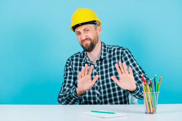 Dissatisfied engineer man is showing stop gesture with hands on blue background