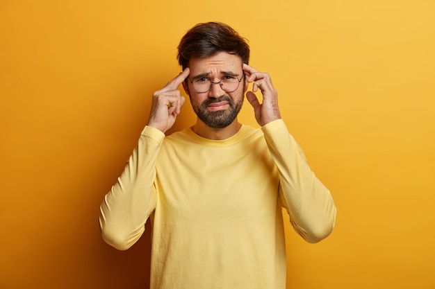 Dissatisfied man thinks intensely, touches temples with index fingers, smirks face, suffers from unbearable headache, wears casual sweater, poses indoor feels pressure and distress, isolated on yellow