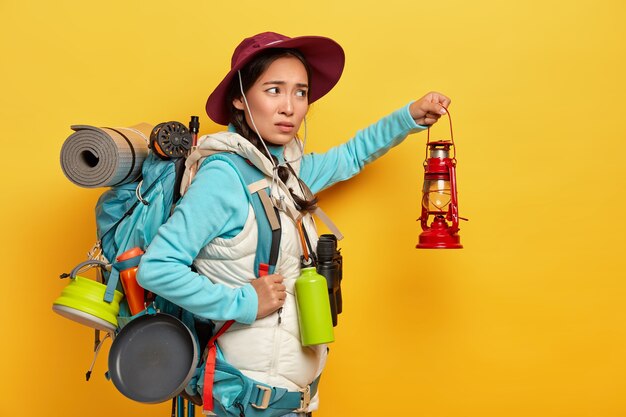 Free photo dissatisfied mixed race female backpacker wears stylish hat and warm vest, holds kerosene lamp to explore surroundings in darkness