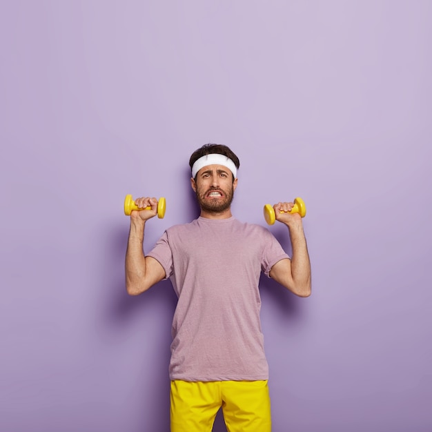 Free photo dissatisfied unshaven man raises arms, trains biceps, wears white headband and active wear, holds dumbbells, has tired look, clenches teeth