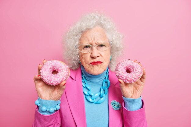 Dissatisfied wrinkled senior woman holds two delicious doughnuts eats junk food wears stylish outfit