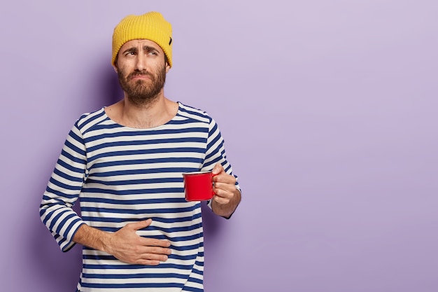 Free Photo dissatisfied young man has stomachache, frowns with discontent, holds cup of hot beverage, wears yellow hat, striped sailor jumper