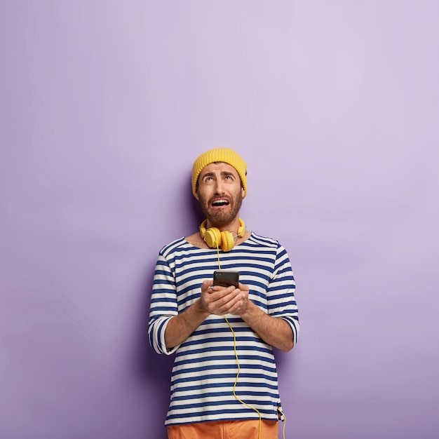 Free photo dissatisfied young man holds modern smartphone, looks above with miserable expression, cannot connect to internet