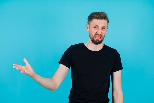 Dissatisfied young man is raising up his hand to left on blue background