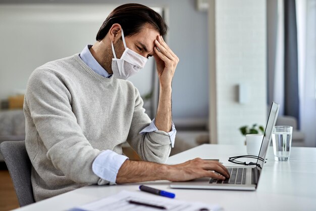 Distraught businessman with protective face mask holding his head in pain while working on a computer at home