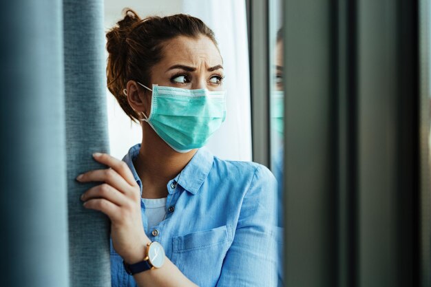 Distraught woman with protective mask on her face looking through the widow at home