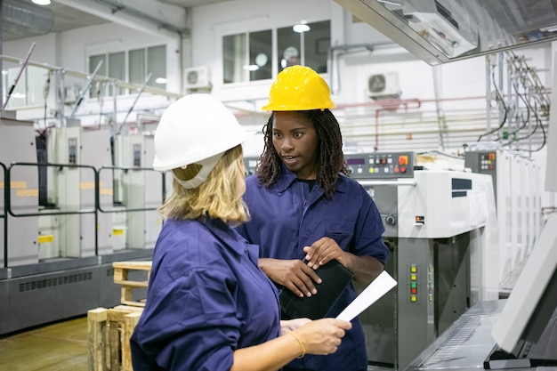 Diverse female factory employees in hardhats and overalls standing on plant floor and chatting