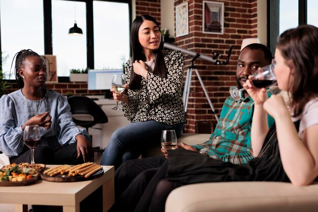 Diverse group of close friends relaxing together at home while hosting wine party in living room. Happy young people enjoying drinking alcohol beverages together while celebrating birthday event.