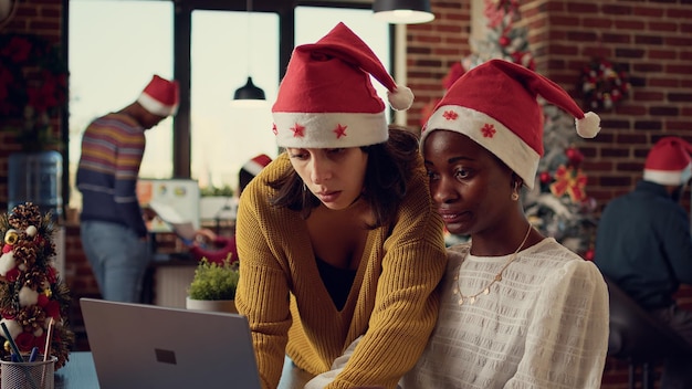 Diverse team of colleagues working on report in festive office, celebrating winter season with christmas tree and decorations. Women looking at laptop and doing teamwork during xmas time.