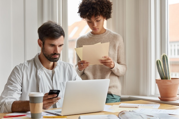 Diverse team of employees have informal meeting at domestic atmosphere, work with paper documents, develop startup during briefing. Focused serious male CEO holds cell phone checks email box on laptop