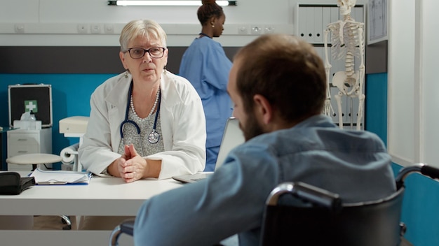 Doctor consulting wheelchair user in disability friendly office to cure chronic health condition. Man with physical impairment attending checkup appointment, receiving advice and support.