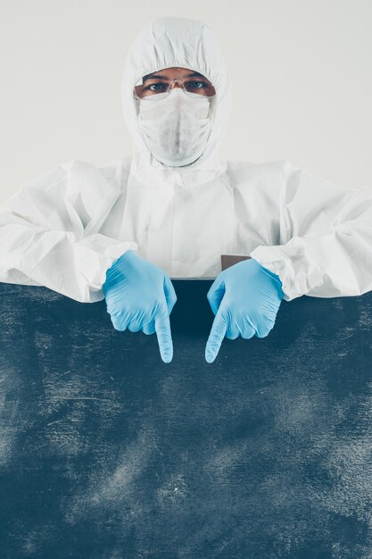 A doctor in mask, gloves and protective suit pointing down and looking in white and dark placard