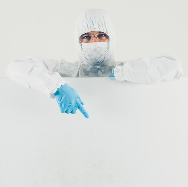 A doctor in mask, gloves and protective suit pointing down in white surface