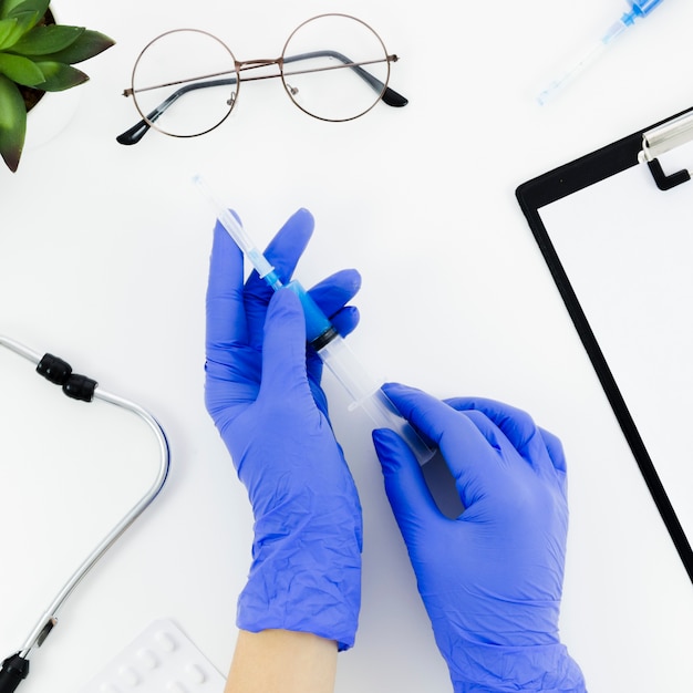 Free photo doctor's hand wearing blue gloves holding syringe on white desk with stethoscope; eyeglasses; pills and clipboard