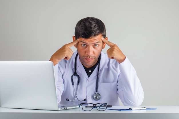 Free photo doctor in white coat, stethoscope touching temples with fingers and looking tired