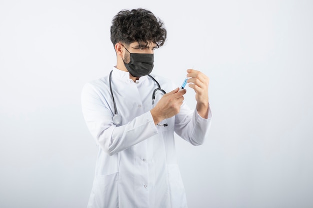 Doctor in white coat with stethoscope holding and touching a syringe isolated on white. 