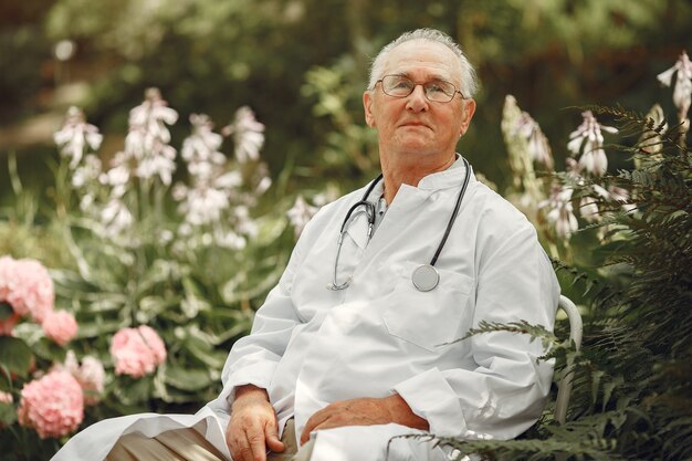 Doctor in a white uniform. Old man sitting in a summer park. Senior with stethoscope.