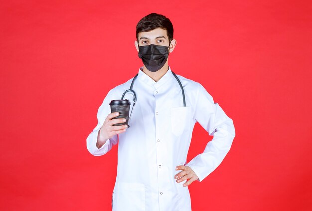 Doctor with stethoscope in black mask holding a black takeaway coffee cup. 