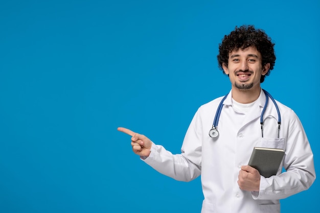 Free photo doctors day curly brunette cute guy in medical uniform smiling and holding a book