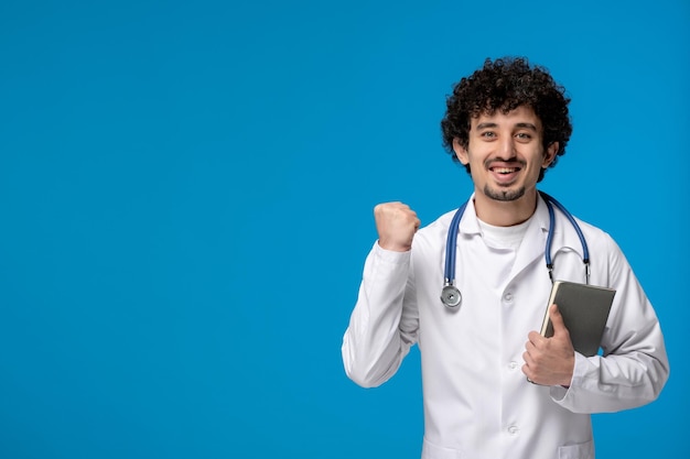 Doctors day curly handsome cute guy in medical uniform holding fist up and a book