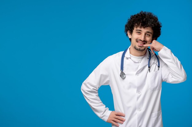 Doctors day curly handsome cute guy in medical uniform making phone call gesture