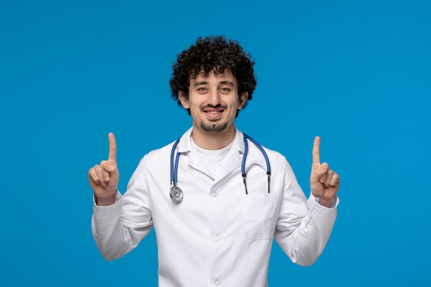 Doctors day curly handsome cute guy in medical uniform pointing up and smiling