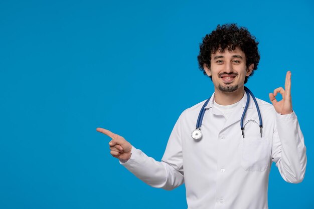 Doctors day curly handsome cute guy in medical uniform showing ok sign gesture