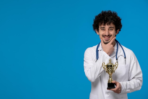 Free photo doctors day curly handsome cute guy in medical uniform smiling and holding a trophy