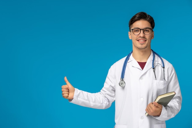 Doctors day cute young brunette guy in lab coat wearing glasses happy holding a book