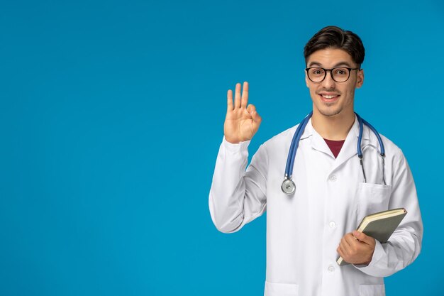 Doctors day cute young brunette guy in lab coat wearing glasses showing ok sign and holding book