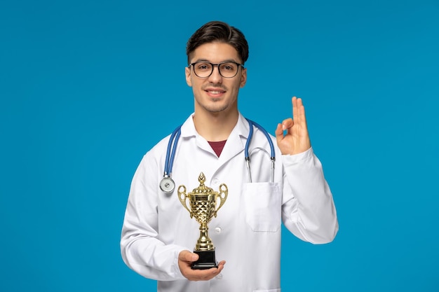 Free photo doctors day cute young handsome man in lab coat and glasses showing ok sign and holding trophy