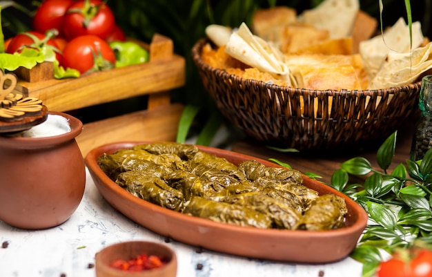 Dolma (tolma, sarma) - stuffed grape leaves with rice and meat. On kitchen table with yogurt, bread, vegetables. Traditional Caucasian, Ottoman, Turkish and Greek cuisine