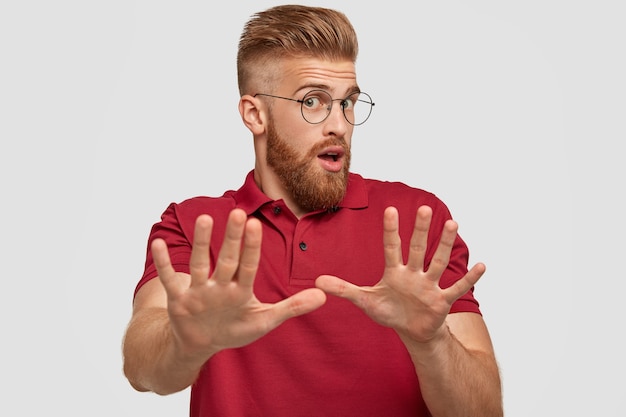 Free photo don`t bother me please! surprised emotional ginger bearded young guy makes stop gesture, stratches hands, feels displeased, wears casual red t-shirt, isolated over white wall.